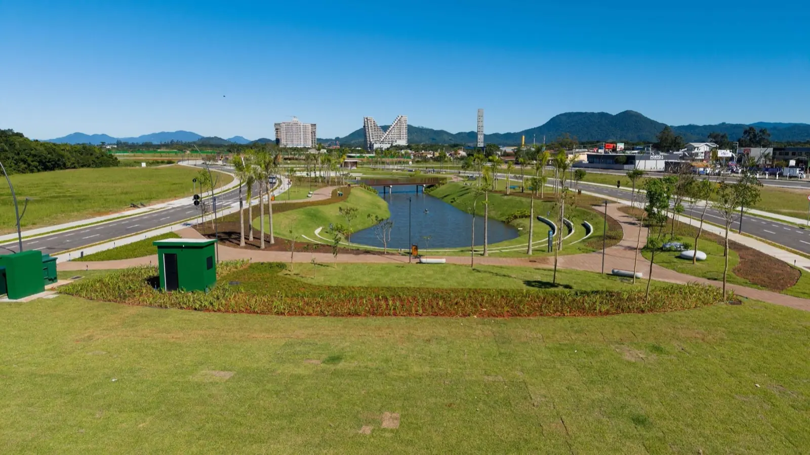 Vista aérea do Parque Viva Equilíbrio: lago, alamedas arborizadas, gramados e vias de acesso, com o skyline de Itapema e montanhas ao fundo